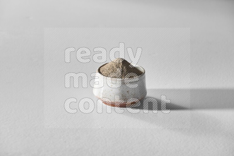 A white ceramic bowl full of black pepper powder and black pepper beads spread on white flooring