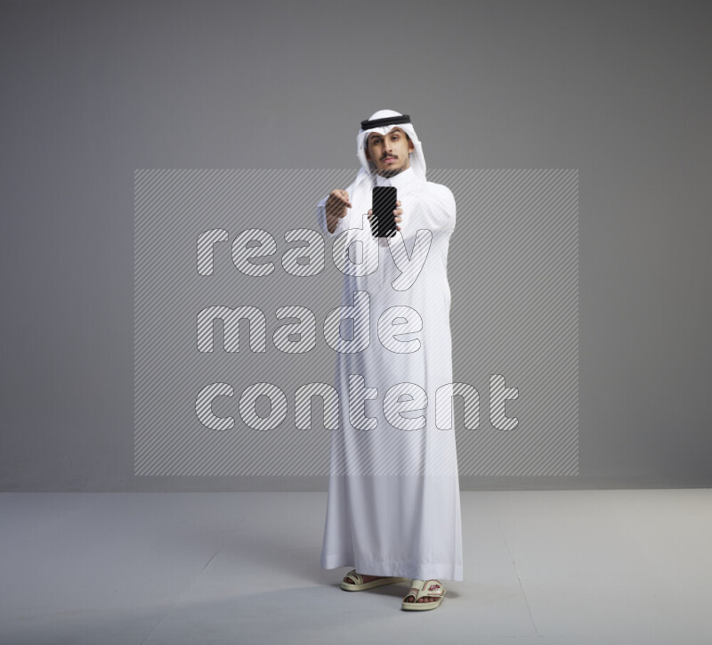 A Saudi man standing wearing thob and white shomag showing phone to camera on gray background