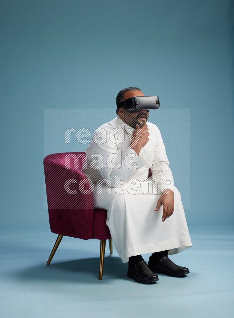 Saudi Man without shimag sitting on chair  gaming with VR on blue background