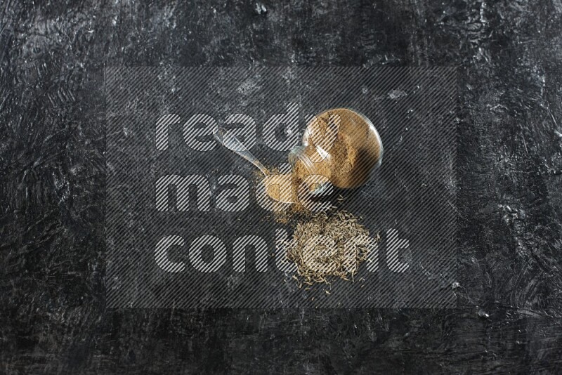 وعاء زجاجي للتوابل مقلوب وملعقة معدنية ممتلئين ببودرة الكمون والبودرة متناثرة مع حبوب الكمون على خلفية سوداء