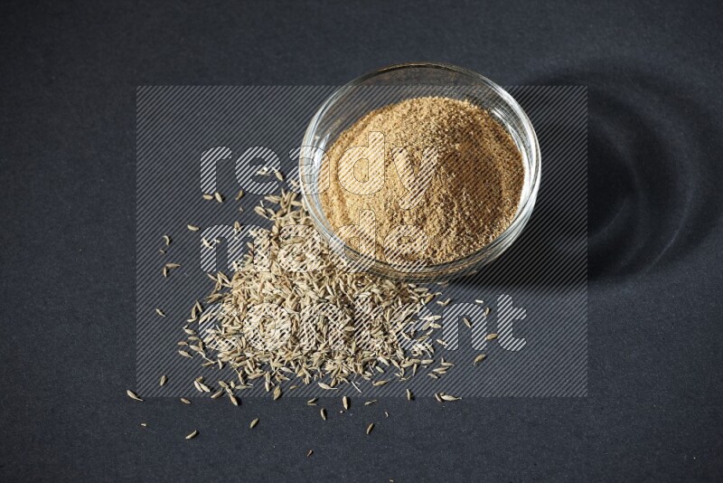 A glass bowl full of cumin powder with cumin seeds beside it on black flooring