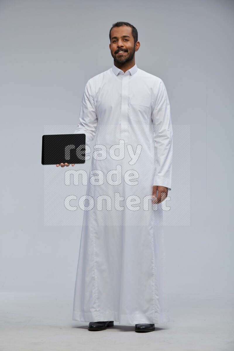 Saudi man wearing Thob standing showing tablet to camera on Gray background