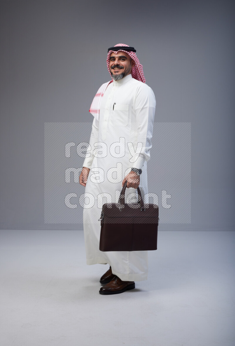 Saudi man Wearing Thob and red Shomag standing holding bag on Gray background