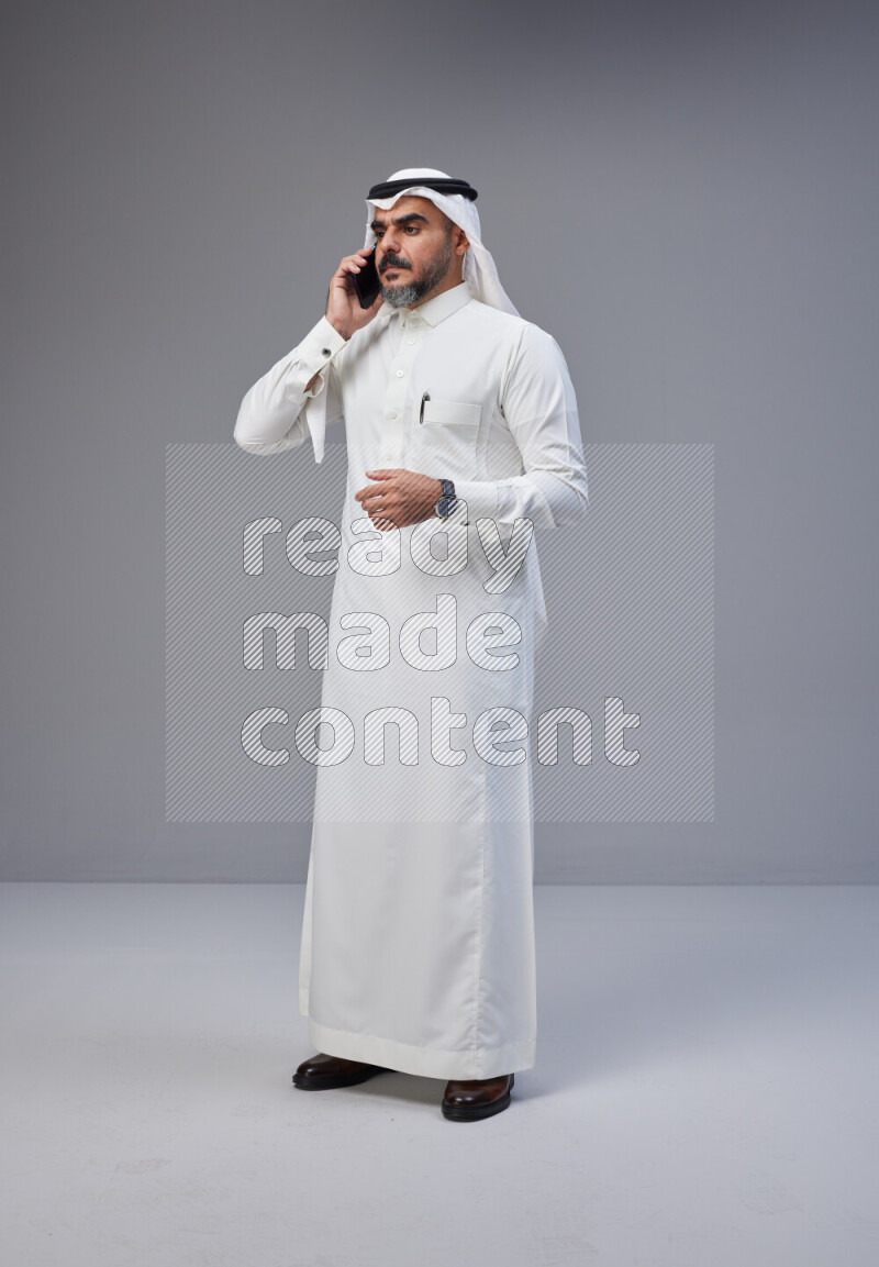 Saudi man Wearing Thob and white Shomag standing talking on phone on Gray background