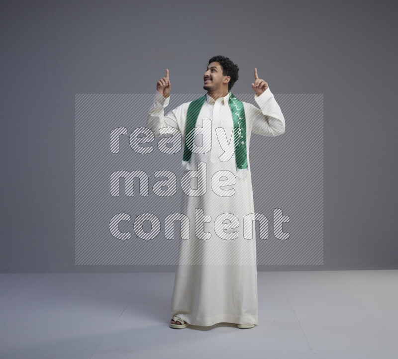 A Saudi man standing wearing thob and Saudi flag scarf on gray background