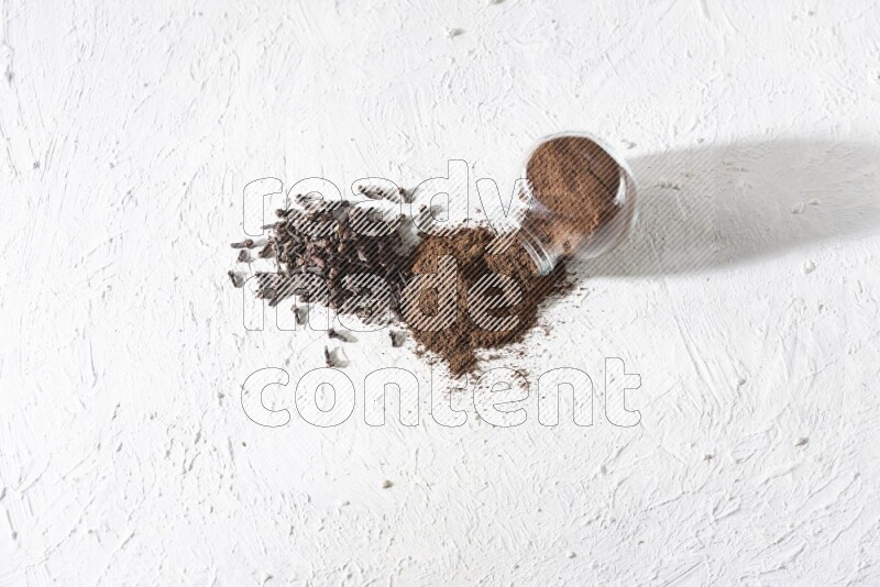 وعاء زجاجي للتوابل مقلوب ممتلئ ببودرة القرنفل وتناثرت البودرة منه مع حبوب القرنفل المنتشرة على أرضية بيضاء