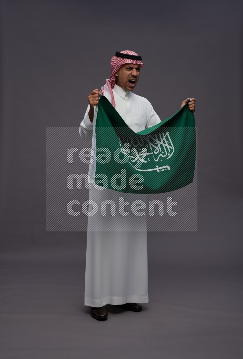 Saudi man wearing thob and shomag standing holding Saudi flag on gray background