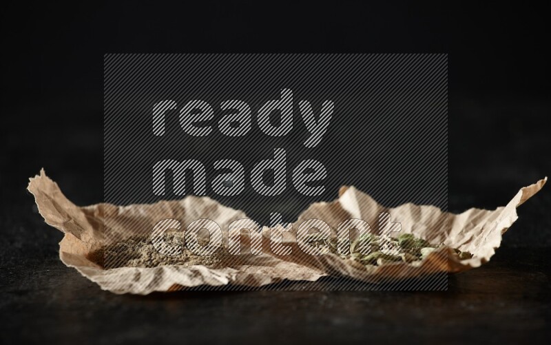 Cardamom seeds and cardamom powder in 2 crumpled pieces of paper on textured black flooring