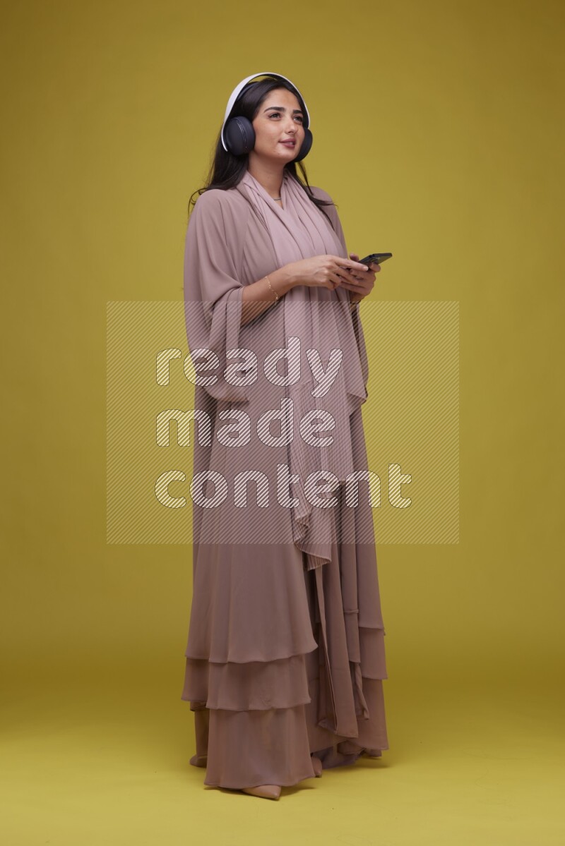 A Saudi woman Listing to Music on a Yellow Background wearing Brown Abaya