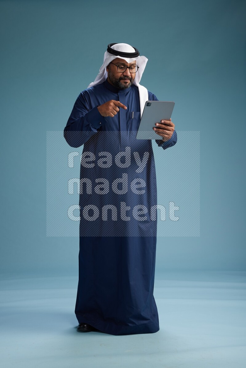Saudi Man with shimag Standing holding ATM card while working on tablet on blue background