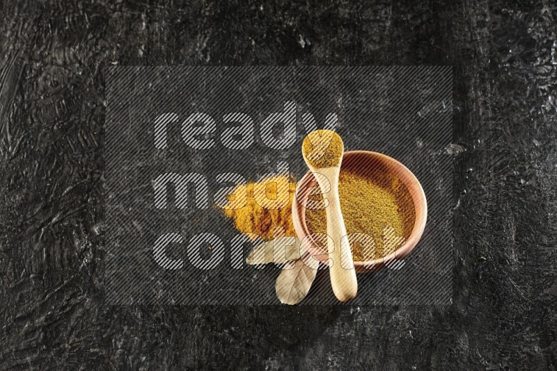 A wooden bowl and a wooden spoon full of turmeric powder on textured black flooring