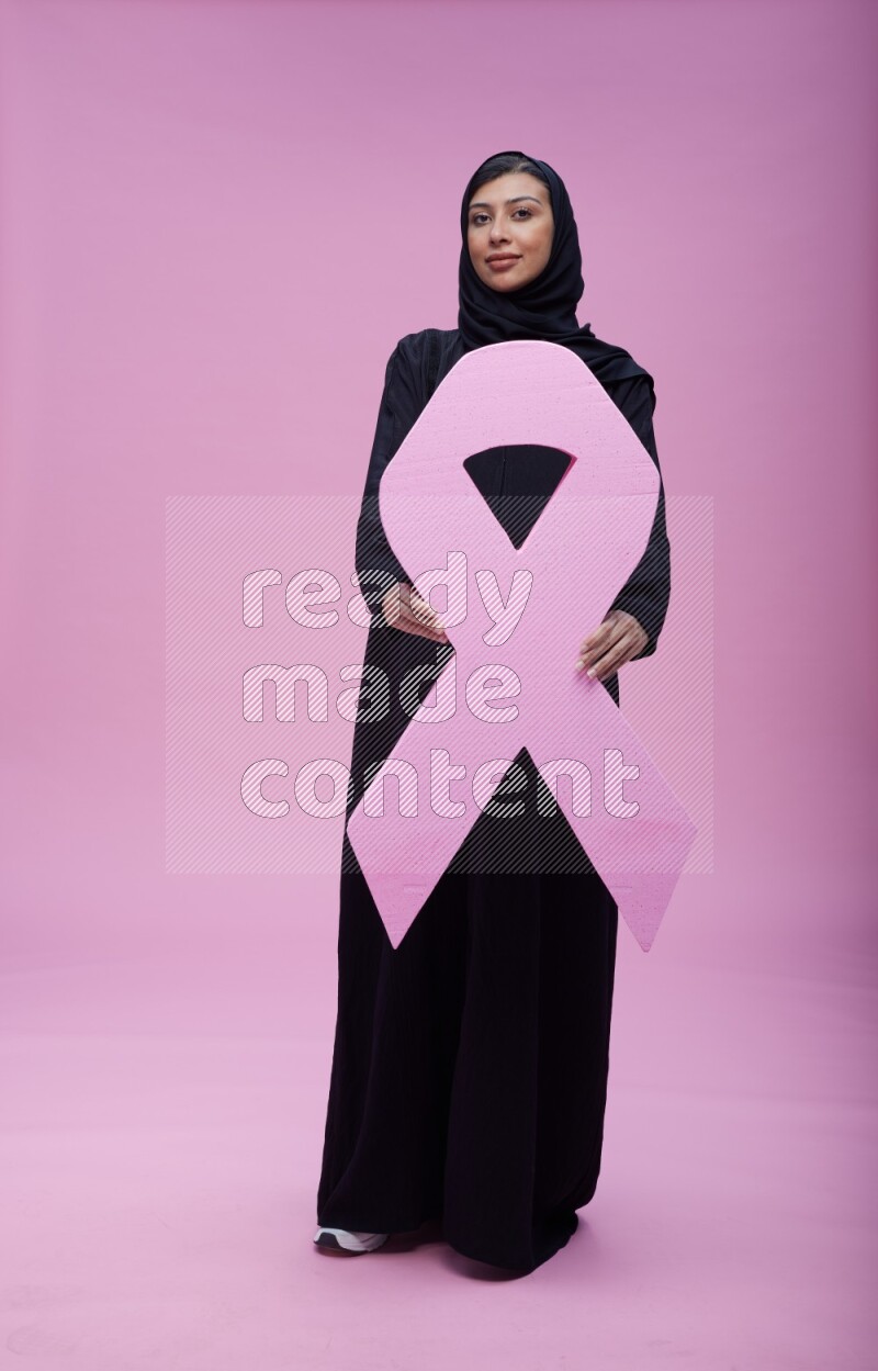 Saudi woman wearing pink ribbon on abaya standing holding awareness ribbon on plain pink background