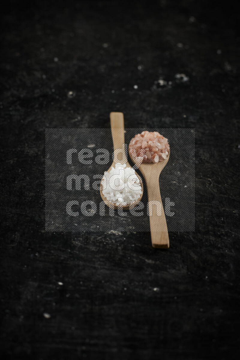 2 wooden spoons filled with fine and coarse salt on black background