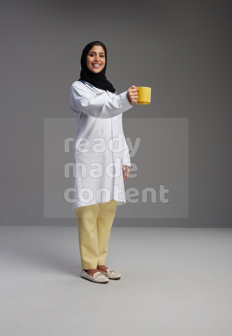 Saudi woman wearing lab coat with stethoscope standing holding mug on Gray background