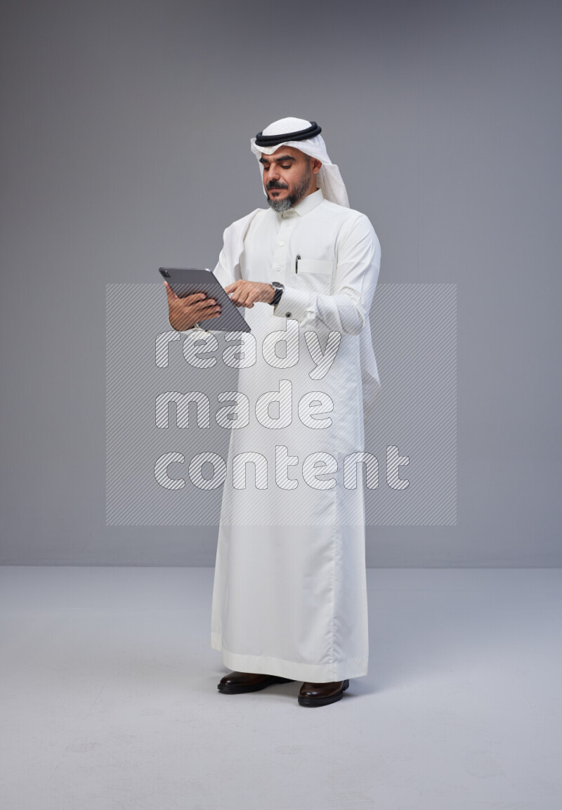 Saudi man Wearing Thob and white Shomag standing working on tablet on Gray background