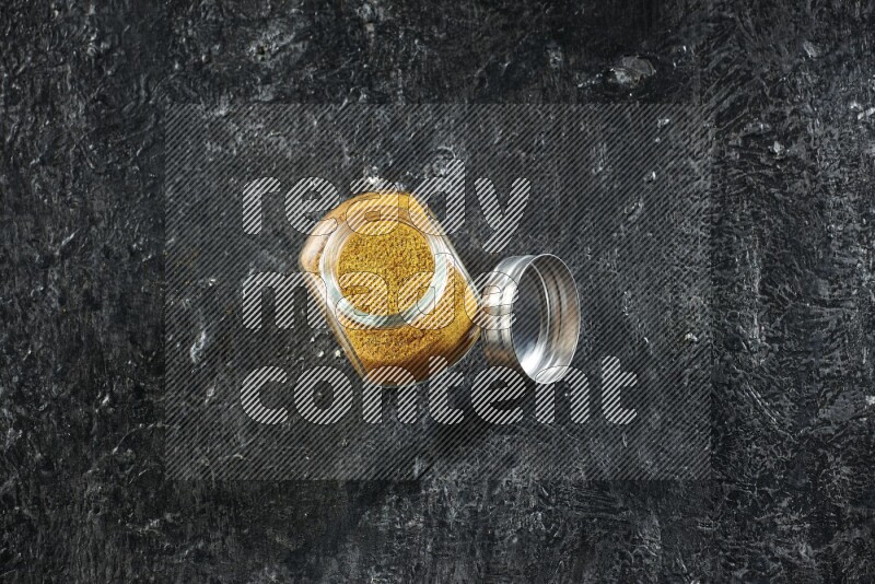 A glass spice jar full of turmeric powder on a textured black flooring