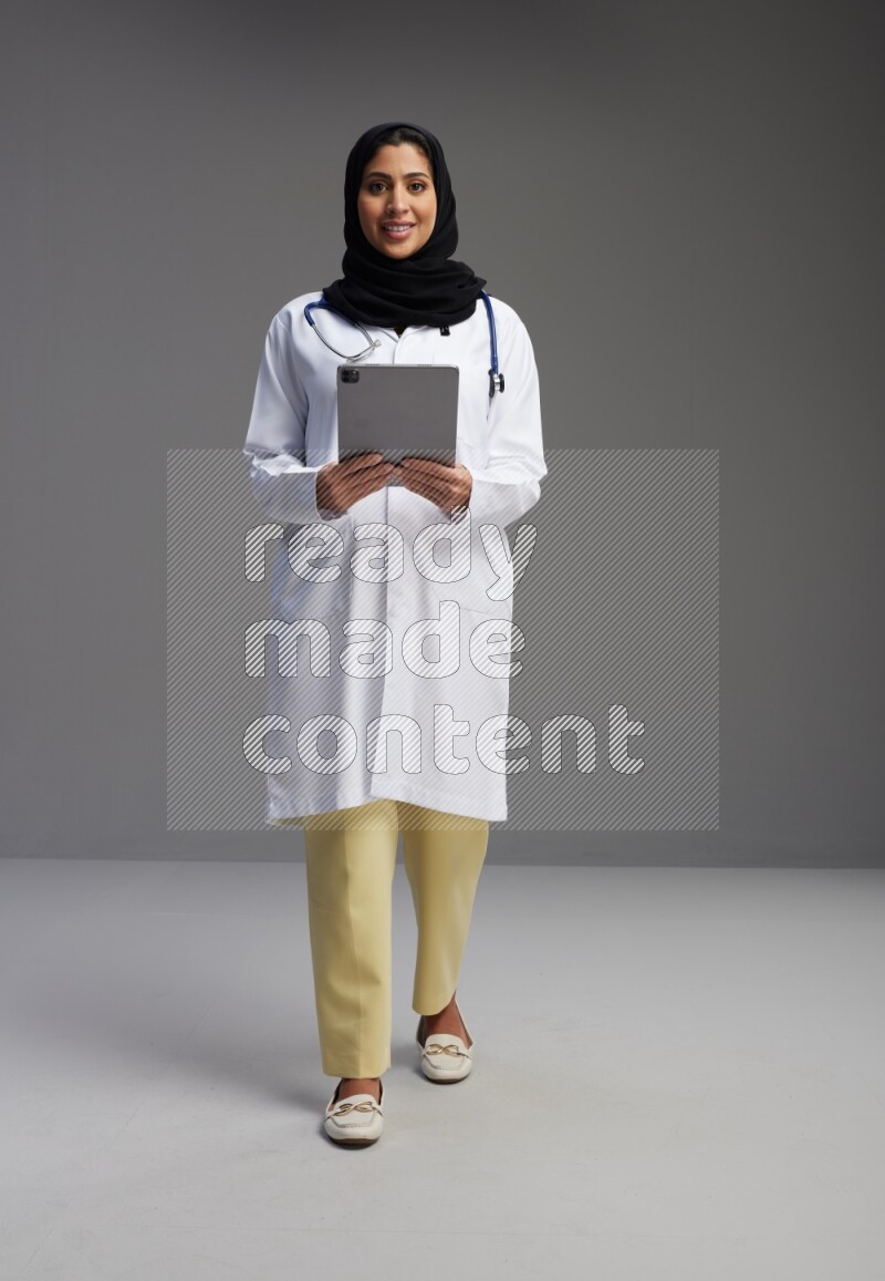 Saudi woman wearing lab coat with stethoscope standing working on tablet on Gray background