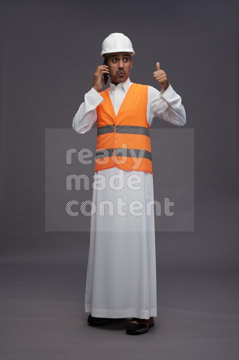 Saudi man wearing thob with engineer vest standing talking on phone on gray background