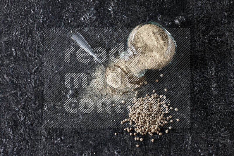 A flipped herbal glass jar and metal spoon full of white pepper powder with spilled powder and pepper beads on textured black flooring