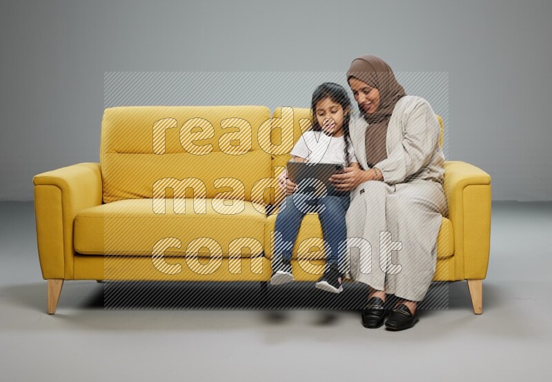 A girl with her mother on yellow sofa and watching on iPad on gray background
