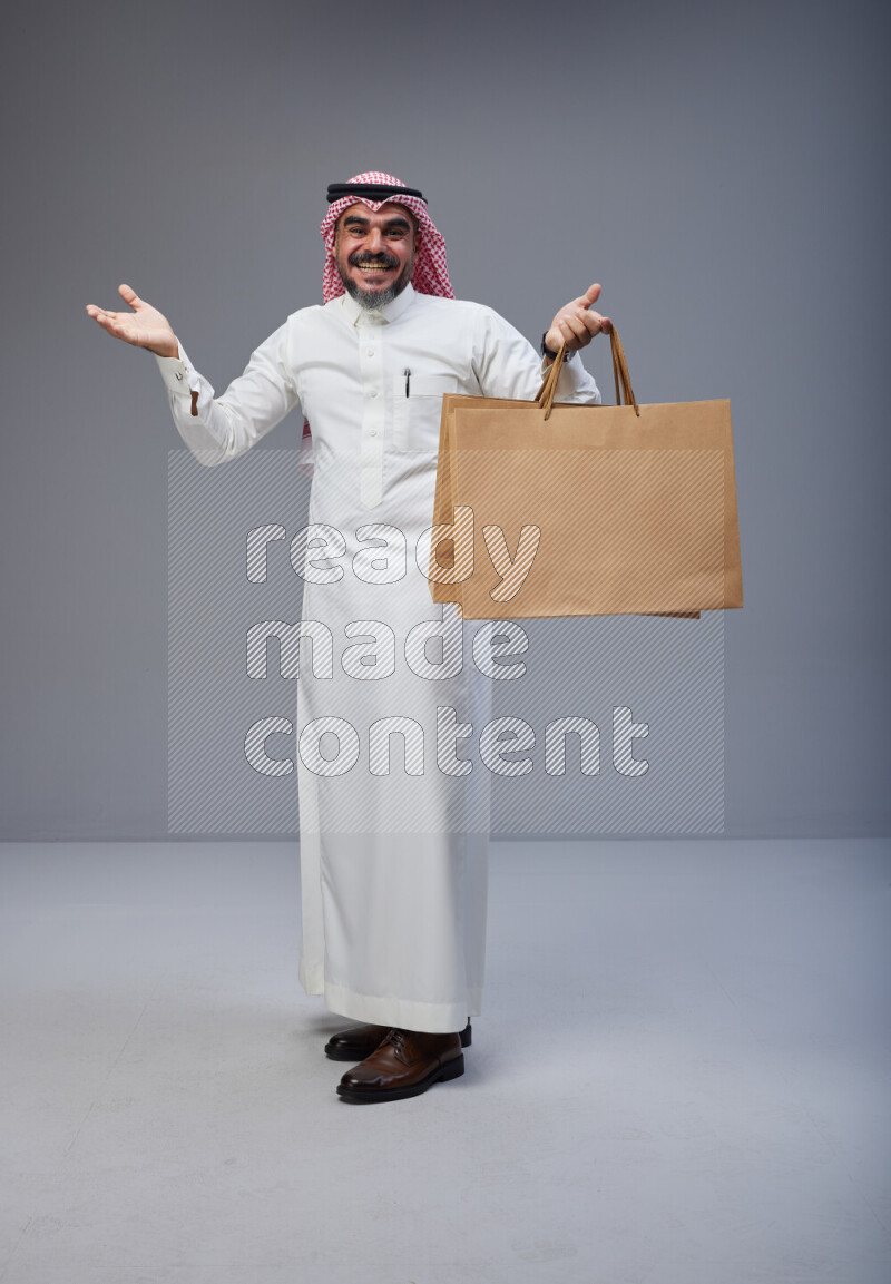 Saudi man Wearing Thob and red Shomag standing holding shopping bag on Gray background