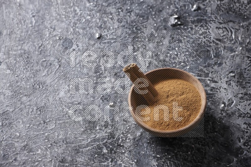 Wooden bowl full of cinnamon powder and a cinnamon stick on a textured black background