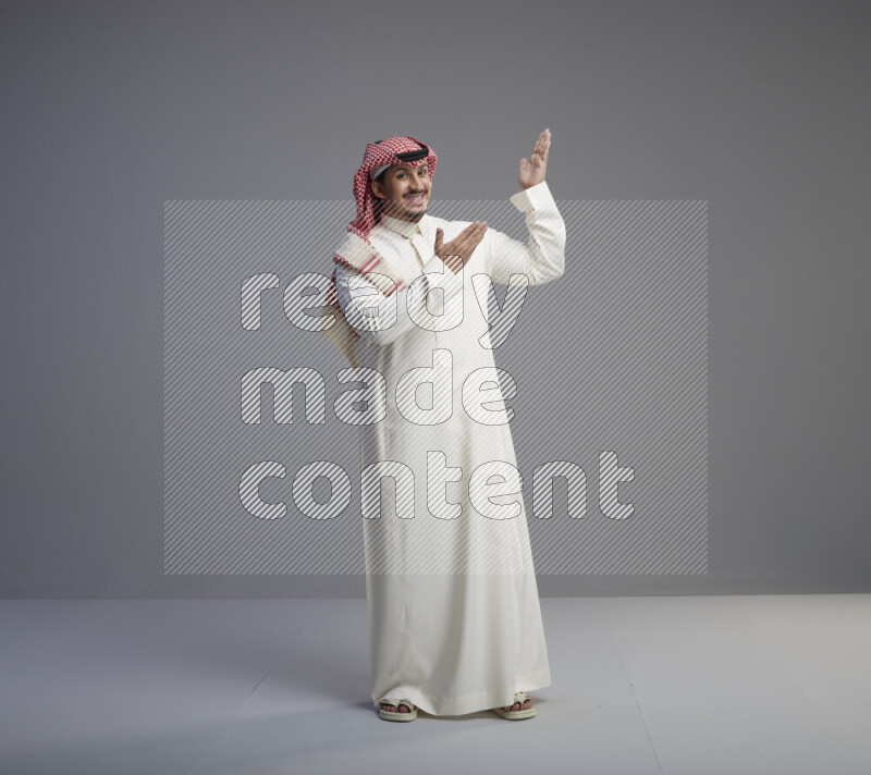 A Saudi man standing wearing thob and red shomag interacting with the camera on gray background