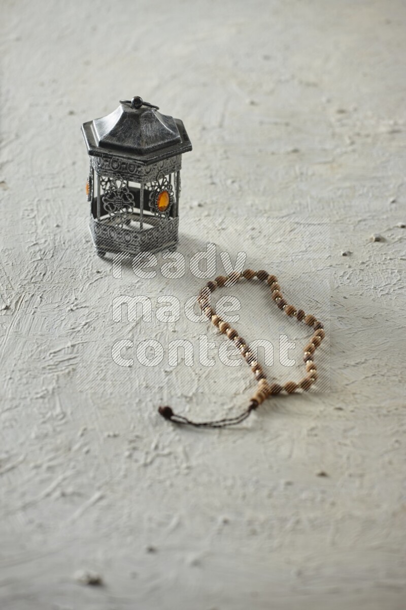 A silver lantern with different drinks, dates, nuts, prayer beads and quran on textured white background