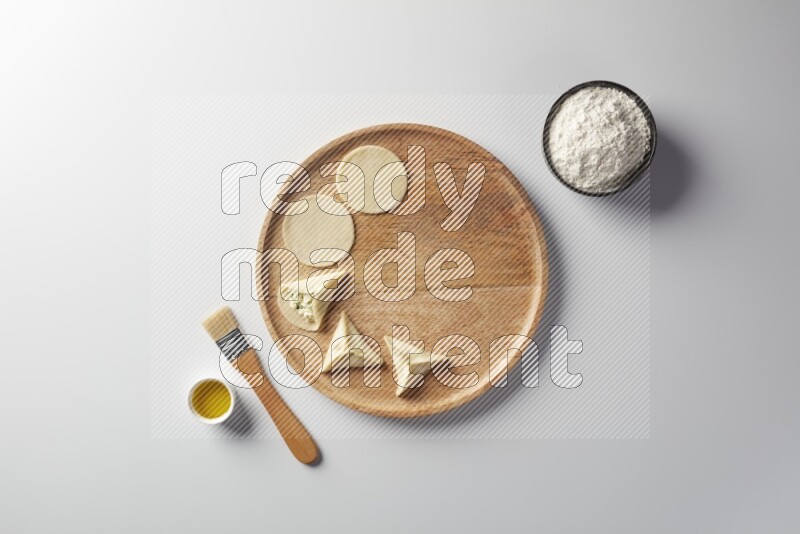 two closed sambosas and one open sambosa filled with cheese while flour, and oil with oil brush aside in a wooden dish on a white background