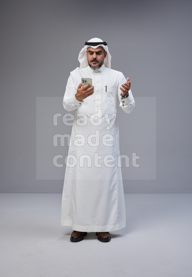 Saudi man Wearing Thob and white Shomag standing texting on phone on Gray background
