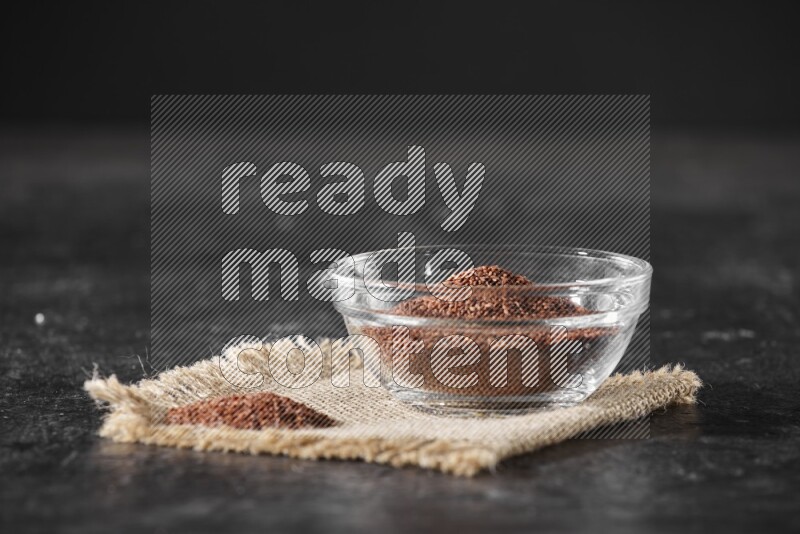 A glass bowl full of garden cress seeds with bunch of the seeds on burlap fabric on a textured black flooring