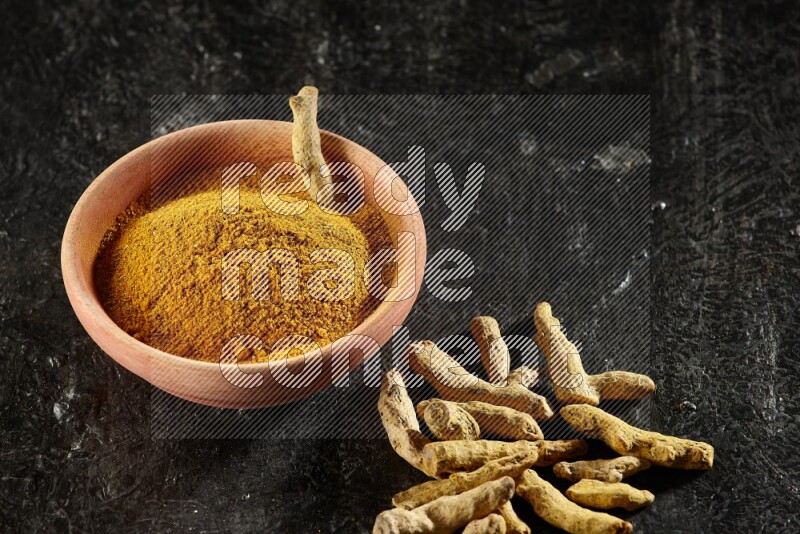 A wooden bowl full of turmeric powder with dried turmeric whole fingers on textured black flooring
