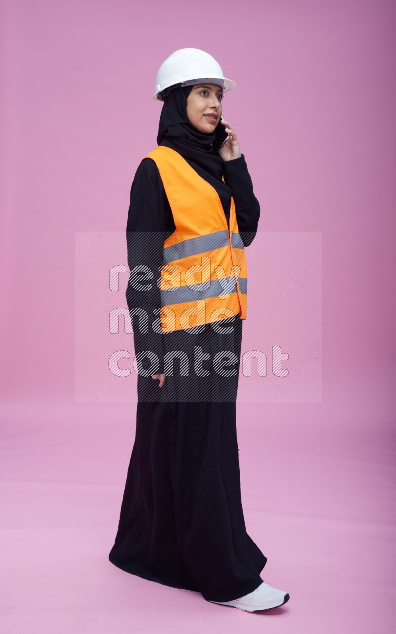 Saudi woman wearing Abaya with engineer vest and helmet standing talking on phone on pink background