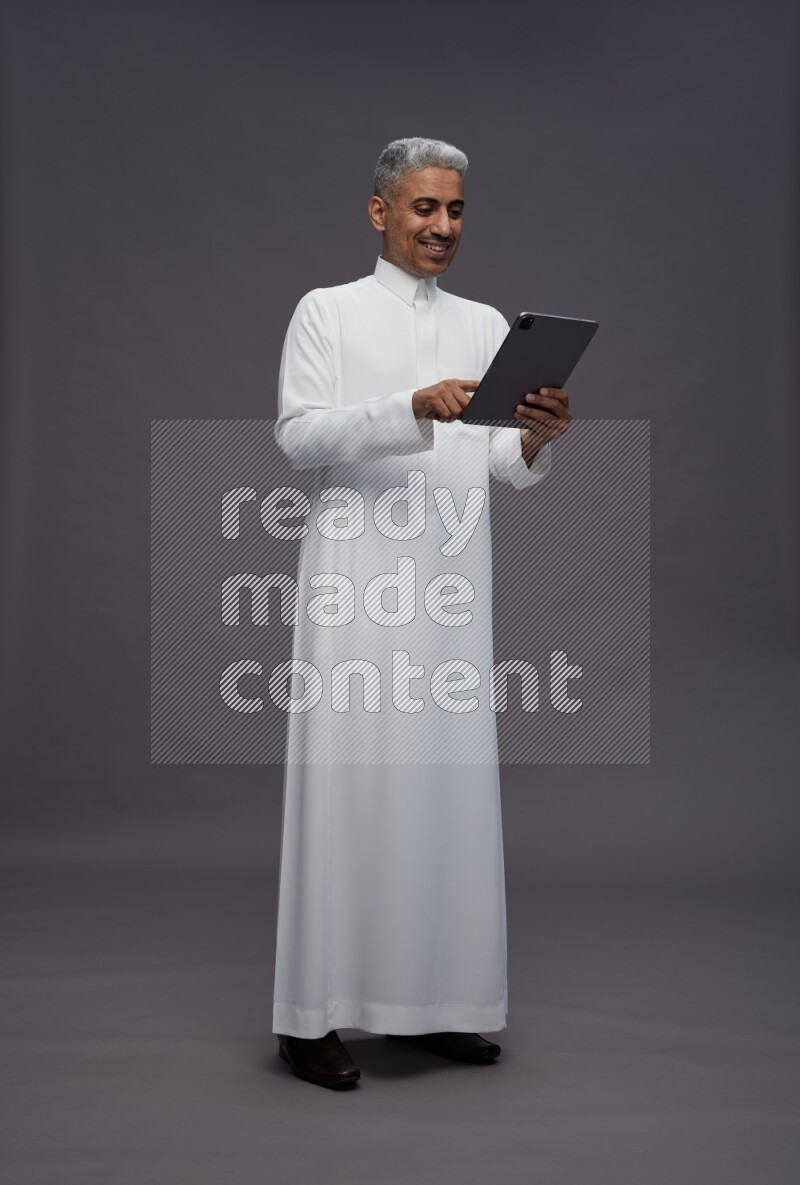 Saudi man wearing thob standing working on tablet on gray background
