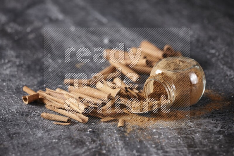 Herbal glass jar full cinnamon powder flipped and a metal spoon full of powder surrounded by cinnamon sticks on textured black background in different angles