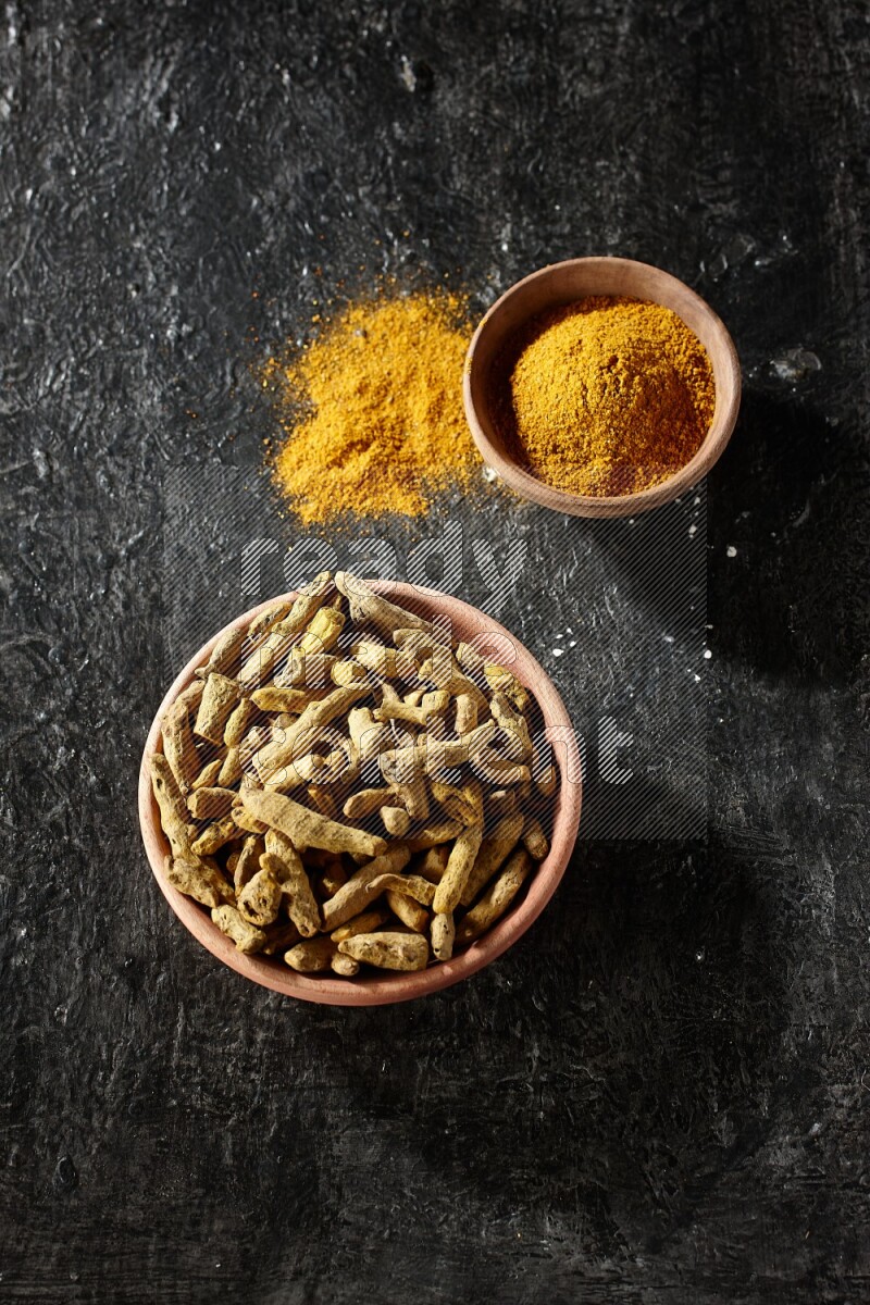 2 wooden bowls, one full of turmeric powder and the other full of dried turmeric whole fingers on a textured black flooring