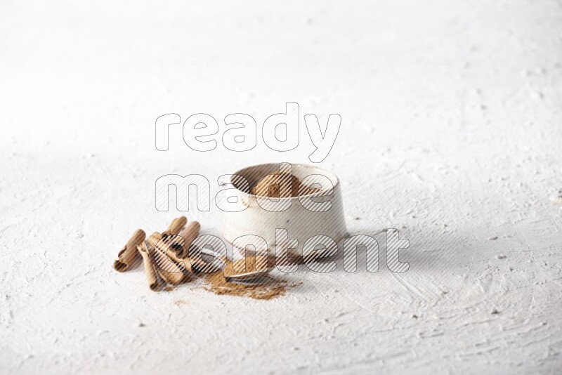 Ceramic beige bowl full of cinnamon powder and a metal spoon with cinnamon sticks next of it on a textured white background