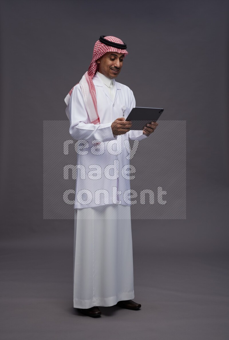 Saudi man wearing thob with lab coat and shomag with pocket employee badge standing working on tablet on gray background