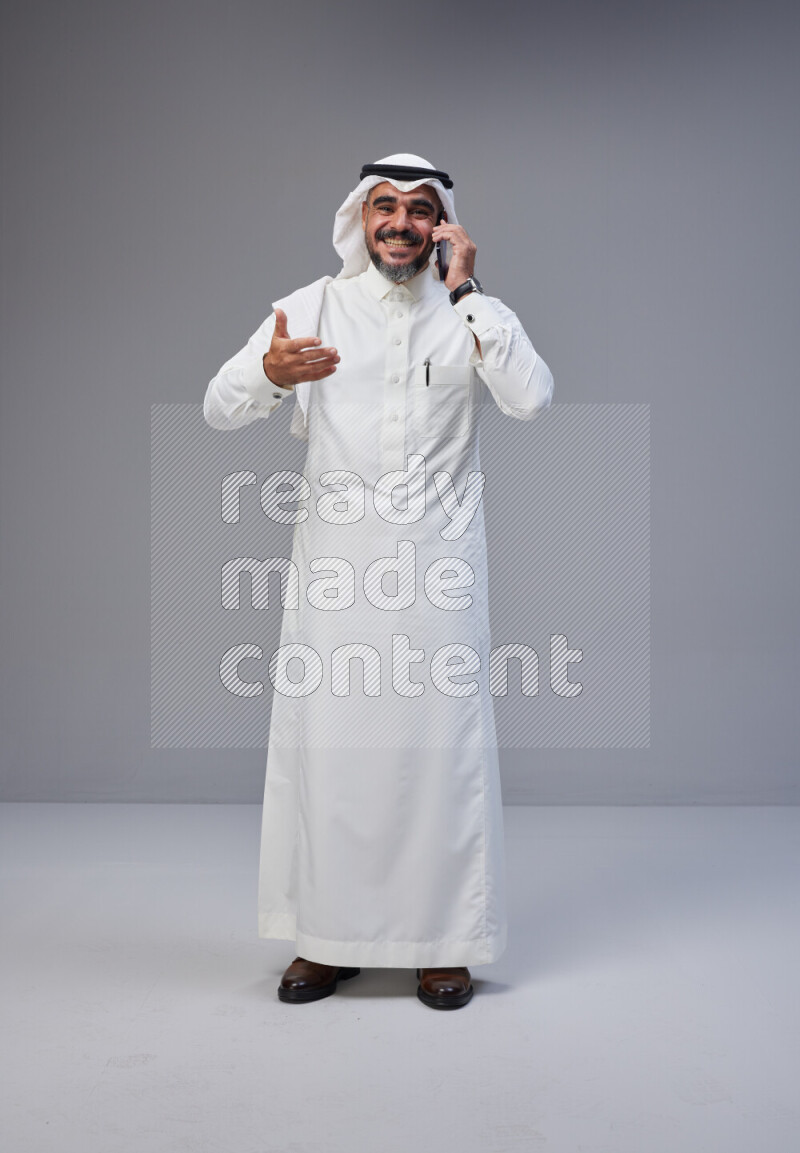 Saudi man Wearing Thob and white Shomag standing talking on phone on Gray background