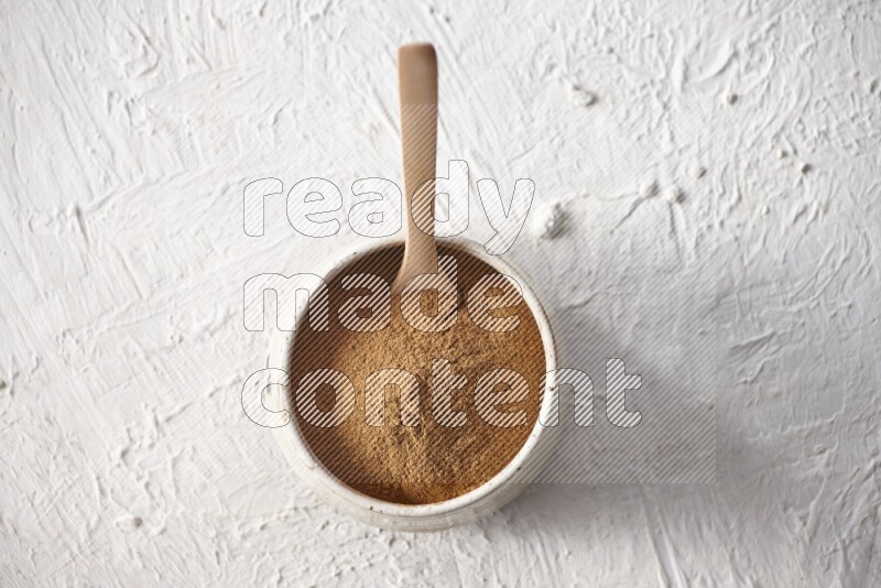 Ceramic beige bowl full of cinnamon powder with a wooden spoon on a textured white background
