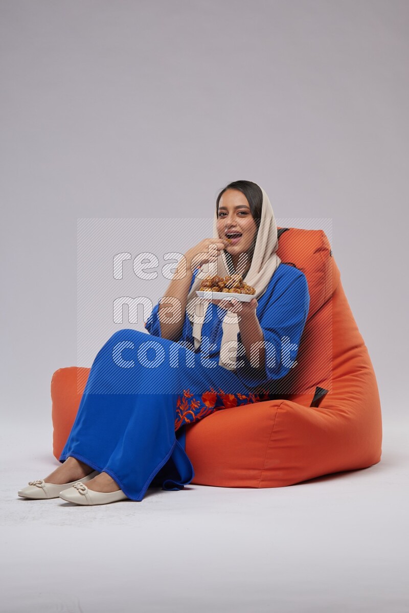 A Woman sitting on an orange beanbag wearing Jalabeya holding a plate of dates