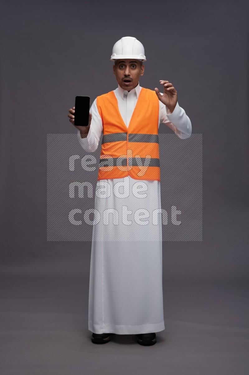 Saudi man wearing thob with engineer vest standing showing phone to camera on gray background