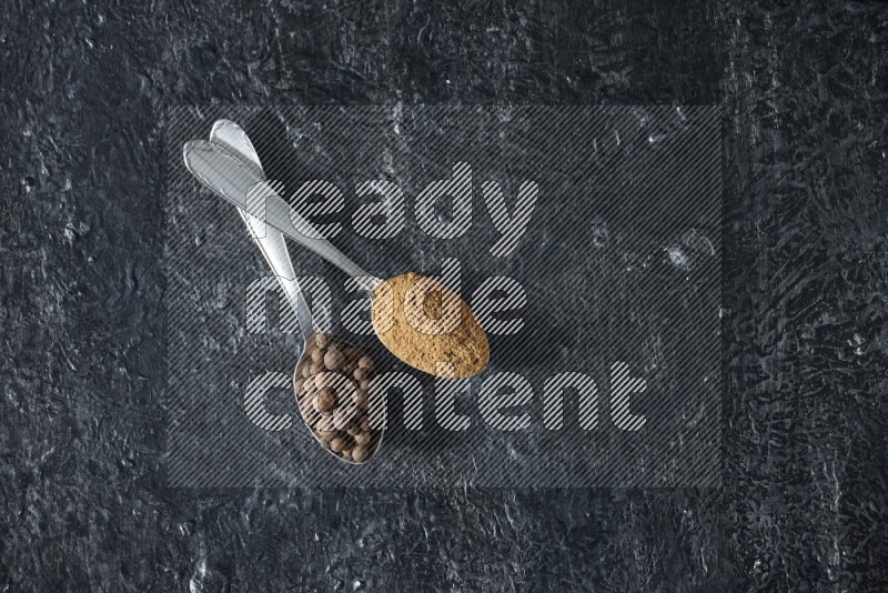 2 metal spoons full of allspice powder and whole balls on a textured black flooring