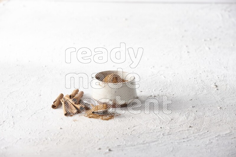 Ceramic beige bowl full of cinnamon powder and a metal spoon with cinnamon sticks next of it on a textured white background