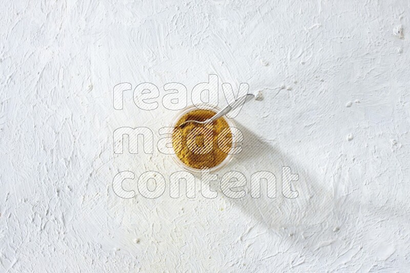 A glass jar and a metal spoon full of turmeric powder on a textured white flooring