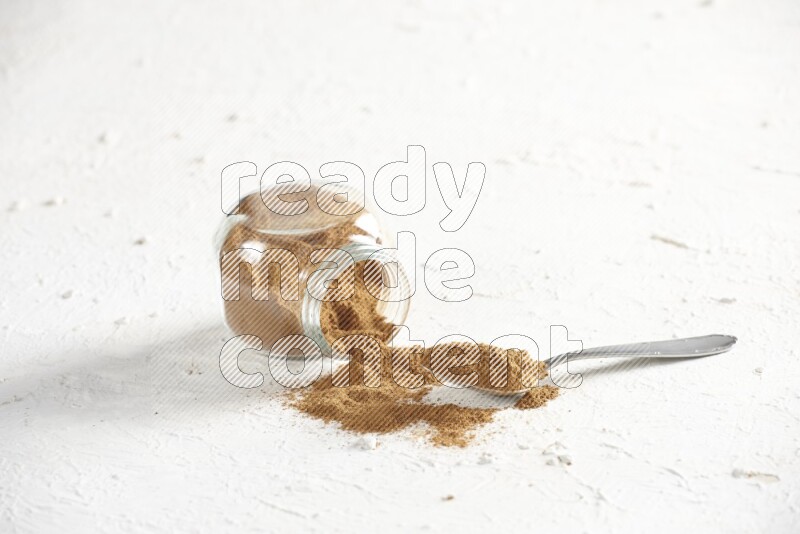 Flipped herbs glass jar full of cinnamon powder with a metal spoon full of powder on a textured white background