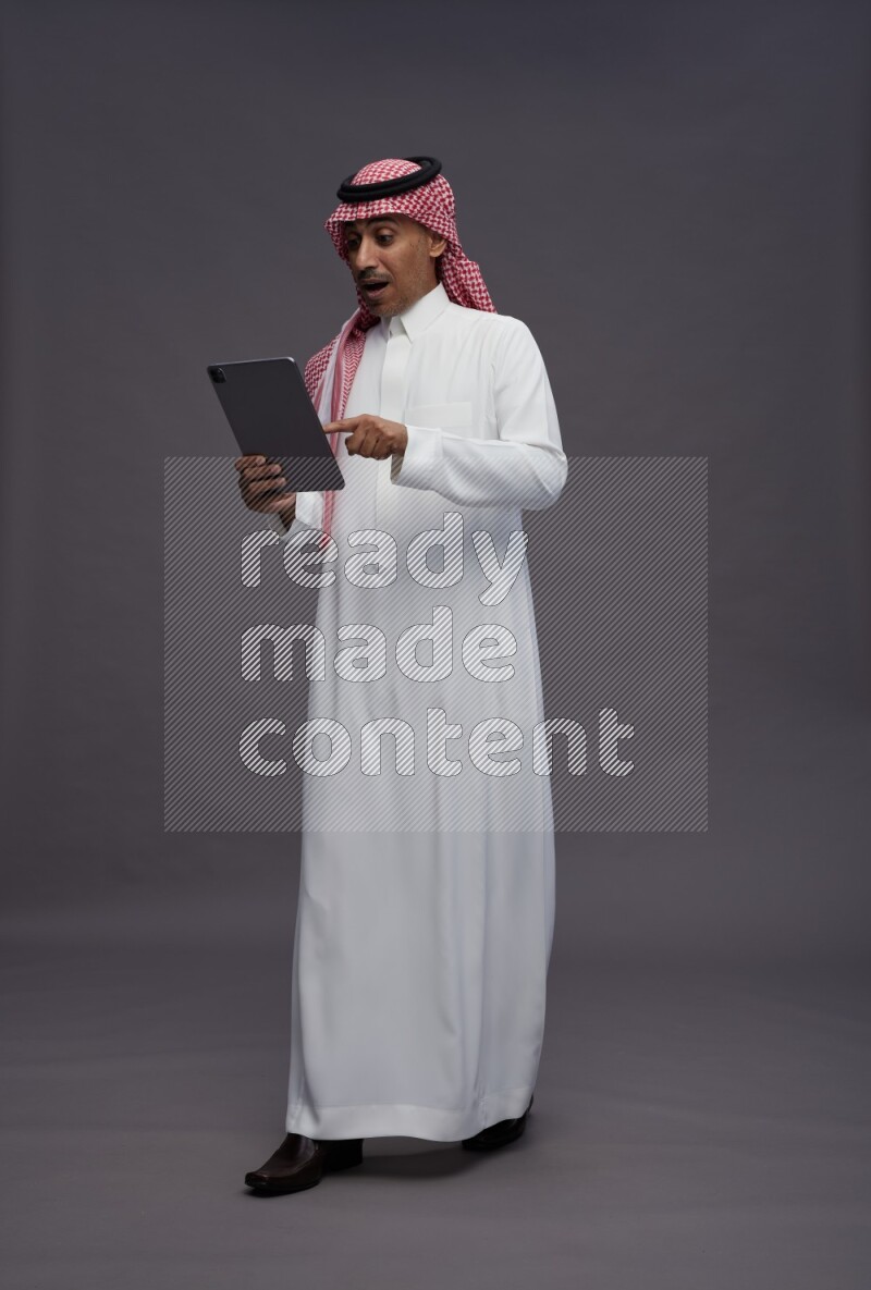 Saudi man wearing thob and shomag standing working on tablet on gray background