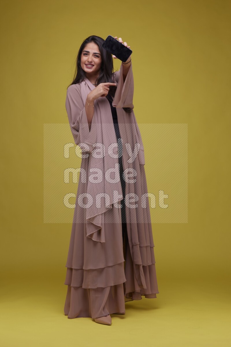 A woman Showing SmartPhone Screen on a Yellow Background wearing Brown Abaya
