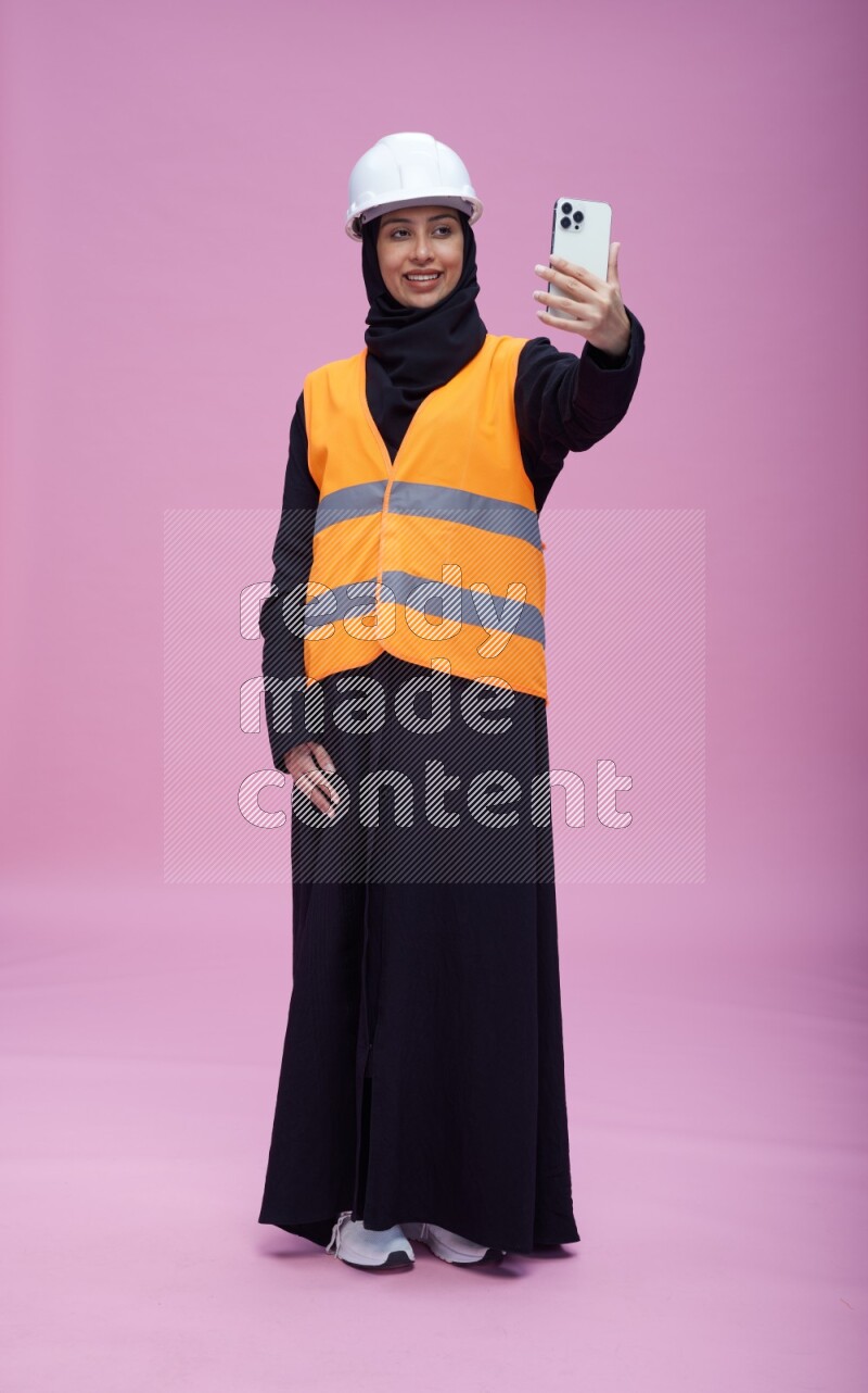 Saudi woman wearing Abaya with engineer vest and helmet standing taking selfie on pink background