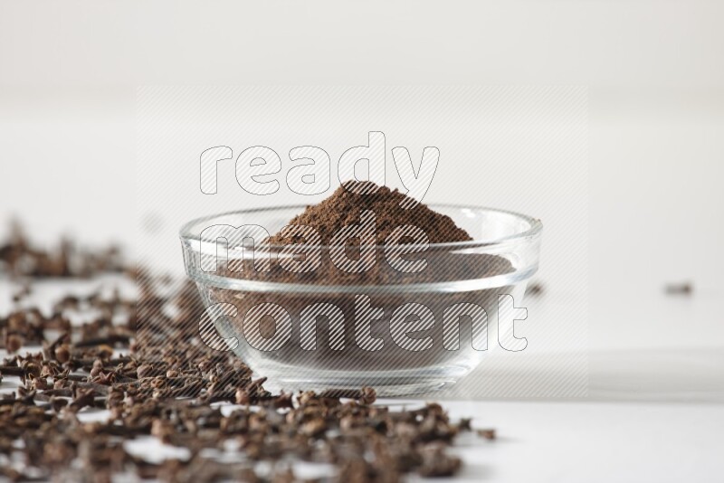 A glass bowl full of cloves powder and cloves grains spread on white flooring